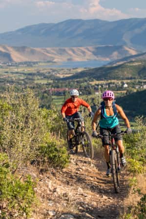 Two bikers making a turn on a mountain trail, with a view of a city in the valley in the background.