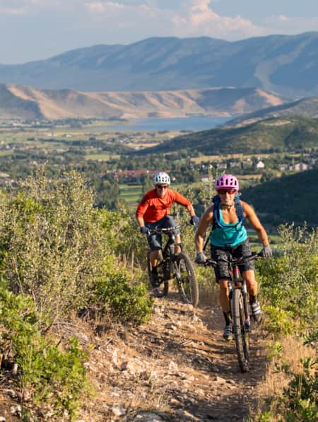 Two bikers making a turn on a mountain trail, with a view of a city in the valley in the background.