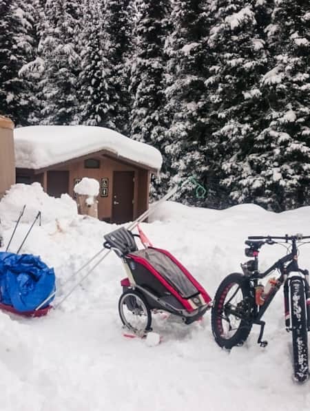 A brown yurt situated by a campsite bathroom, surrounded by snow, with a mountain bike in front of the yurt.