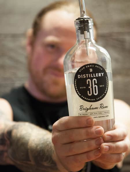 A man holding the bottom of a glass bottle with both hands, with a label that reads 'Rum 36 Brighman Rum.'