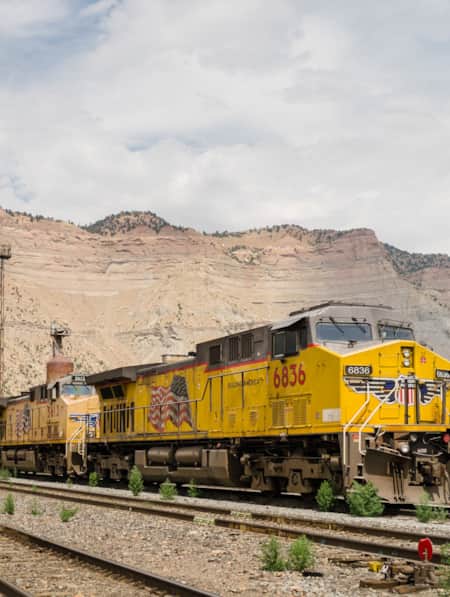 A yellow train arriving in a small town with a large cliff face in the backdrop.