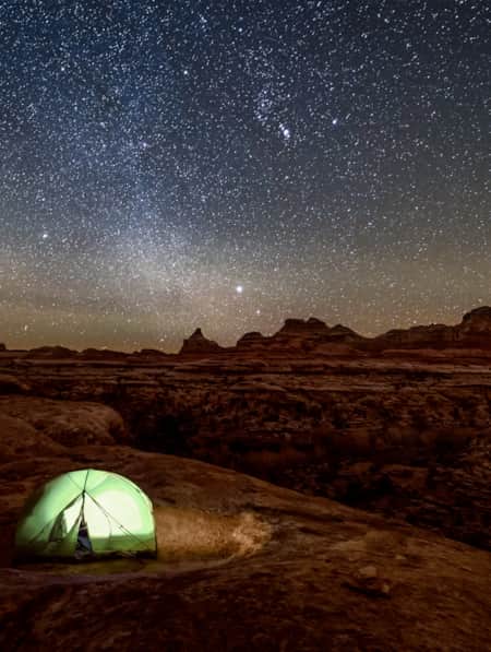An expansive view of a red rock desert illuminated by the night sky, with a green tent in the landscape and a light on inside the tent.