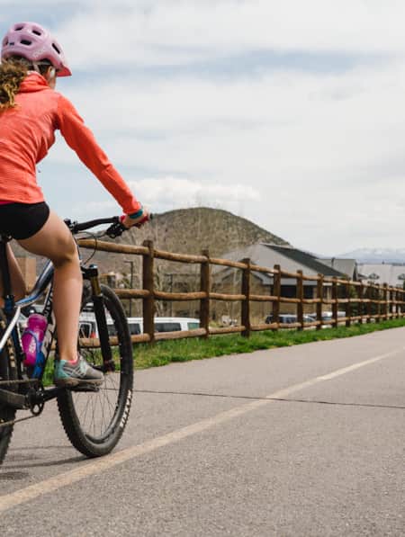 A young girl wearing a helmet biking on a paved walking trail through a small town.