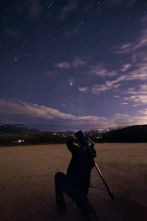 A person kneeling and looking through a telescope at a star-filled night sky.