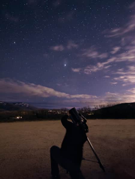 A person kneeling and looking through a telescope at a star-filled night sky.