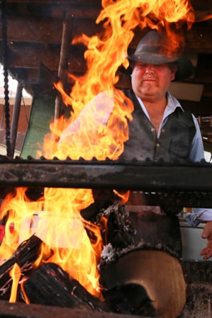 A man in a cowboy hat stands behind a grill with large flames, keeping them under control.