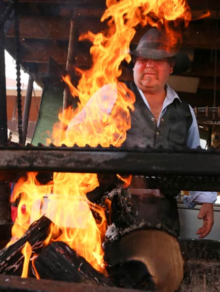 A man in a cowboy hat stands behind a grill with large flames, keeping them under control.