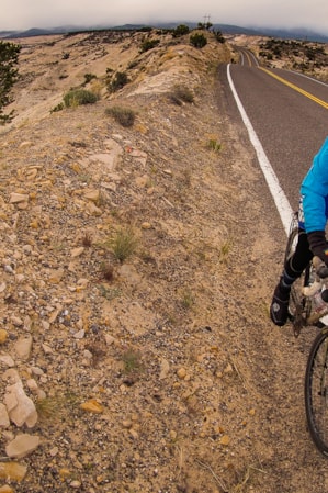 A person cycling along the side of a road through a landscape of large, rocky hills.