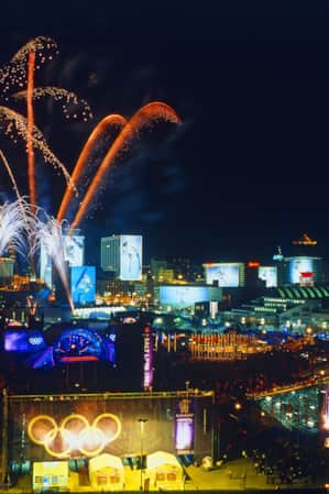 Fireworks lighting up the night sky at a plaza during the Olympics, with stadium seating and screens displaying Olympic athletes.