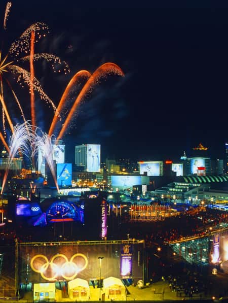 Fireworks lighting up the night sky at a plaza during the Olympics, with stadium seating and screens displaying Olympic athletes.