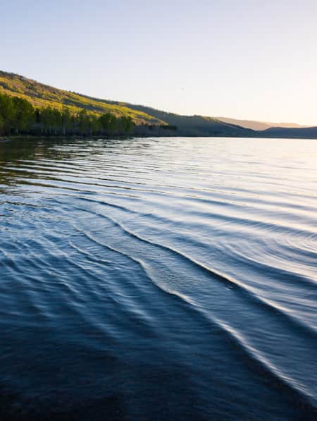Landscape view of a lake with green mountains covered in trees and bushes on the left side, and the sun gleaming over the top of a mountain on the horizon to the right.
