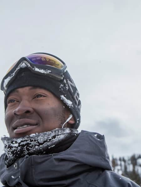 A man standing outdoors with mountains in the background, smiling as he gazes into the distance, dressed warmly in winter clothes.