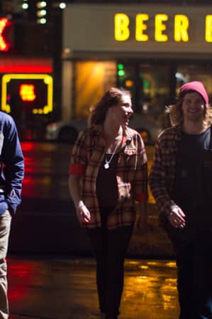 A group of young people walking at night on a city street, with a neon sign reading 'Beer Bar' behind them.