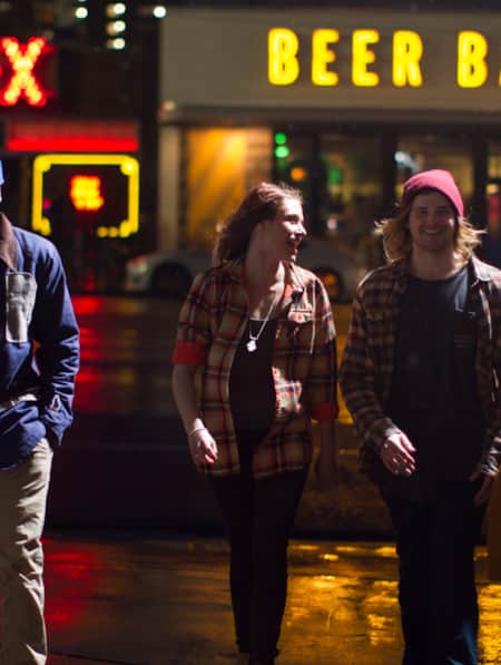A group of young people walking at night on a city street, with a neon sign reading 'Beer Bar' behind them.