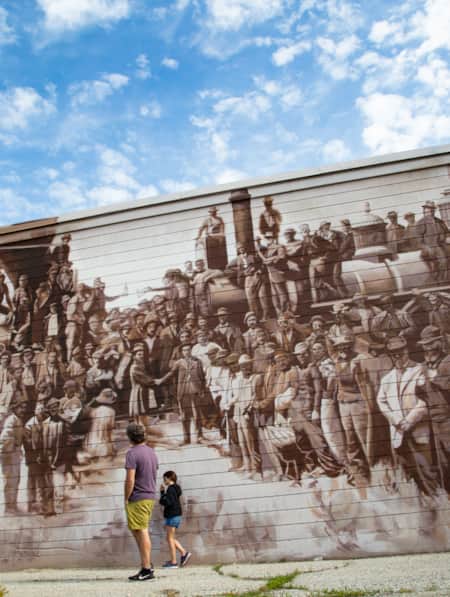 A man and a young girl admiring a large mural depicting an old-time scene of people at a railroad.