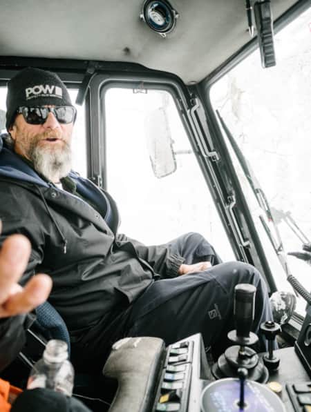 A man sitting in the driver's seat of a snowcat, with his arm extended out, welcoming someone.
