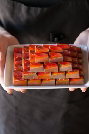 A person holding a white rectangular plate of chocolates, with each morsel adorned with an orange decorative glaze.