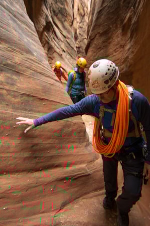 Three people walking through a narrow slot canyon, wearing helmets and carrying rope.