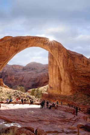 A natural bridge shaped like an arch, made of red sandstone, with hikers gathered at its base.