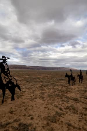 Metal sculptures of horses and cowboys in an open grass field with mountains in the background.
