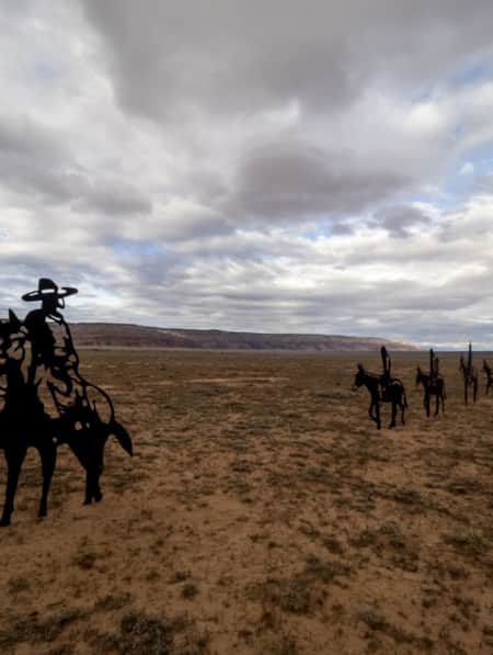 Metal sculptures of horses and cowboys in an open grass field with mountains in the background.