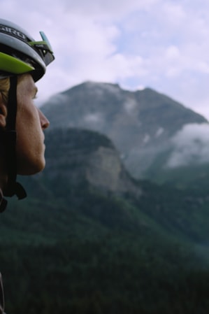 A man wearing a biking helmet, looking off into the distance with foggy, lush green mountains in the background.