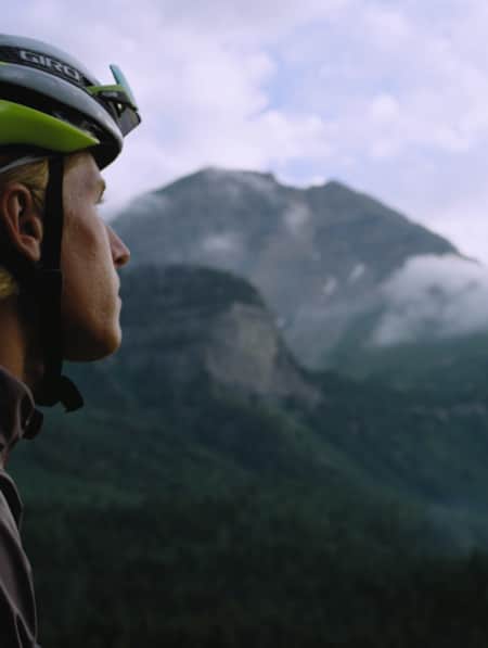 A man wearing a biking helmet, looking off into the distance with foggy, lush green mountains in the background.