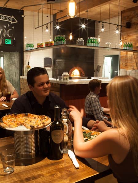 Two people sharing a pizza and bottle of wine, holding hands and looking at each other across the table.