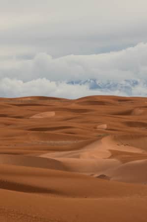 Orange sand dunes in front of a cloudy mountain range with snow.