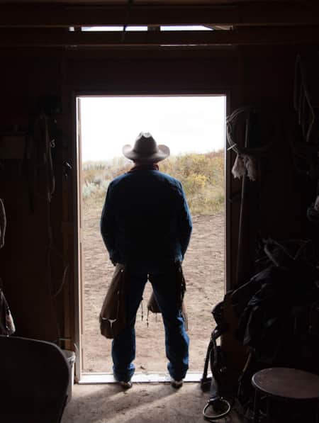 A man standing at the entrance of a barn, wearing chaps and a cowboy hat.