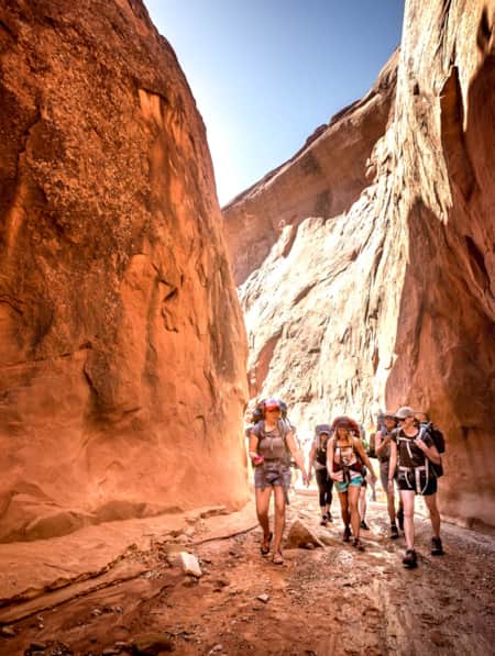 A group of women with backpacks hiking through a spacious slot canyon.