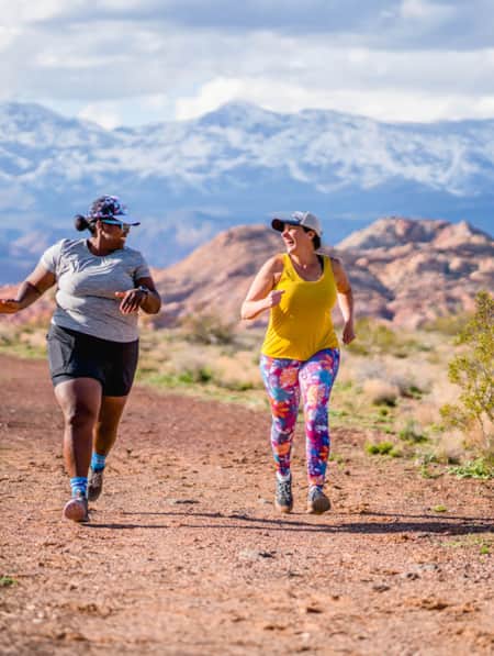 Two women running on a dirt trail, surrounded by open fields, with a mountain range visible in the background.