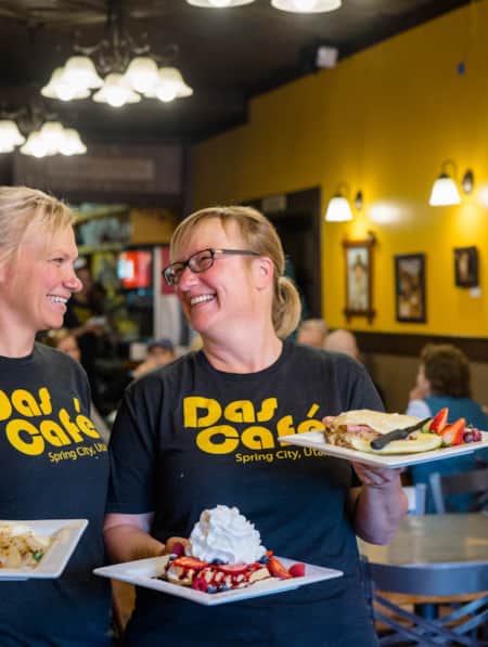 Two women smiling and looking at each other while standing in a restaurant, each holding a plate of breakfast foods.