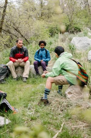 A group of children and adults sitting in a circle on rocks in the woods, engaging in conversation with one another.