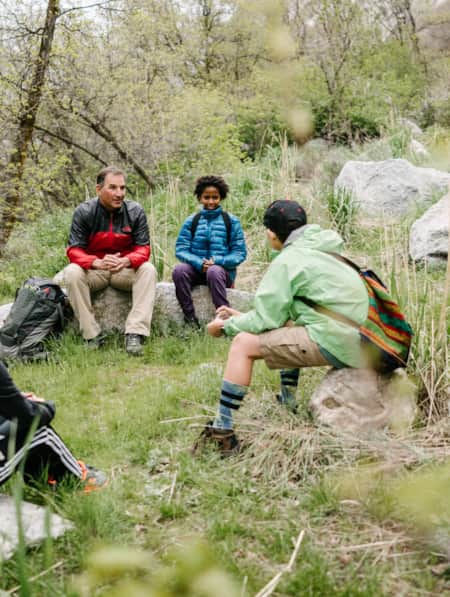 A group of children and adults sitting in a circle on rocks in the woods, engaging in conversation with one another.