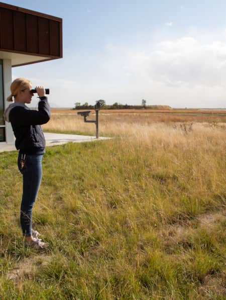 A woman standing outside a building overlooking a grassy field, and using binoculars to look into the distance.