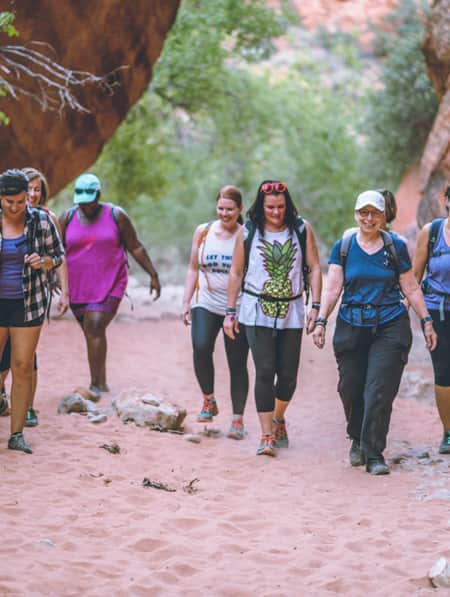 A group of women hiking on sand through the desert, with large red rocks on either side.