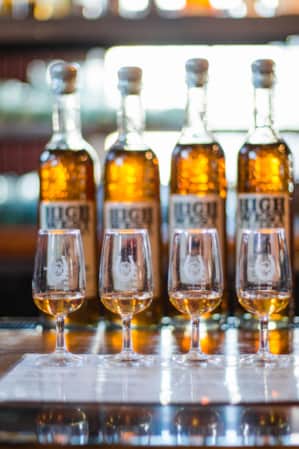 A row of tasting glasses, each with a small amount of whiskey, with whiskey bottles displayed behind them.