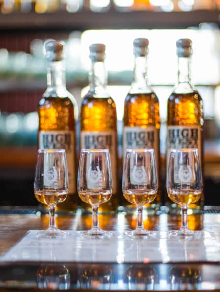 A row of tasting glasses, each with a small amount of whiskey, with whiskey bottles displayed behind them.