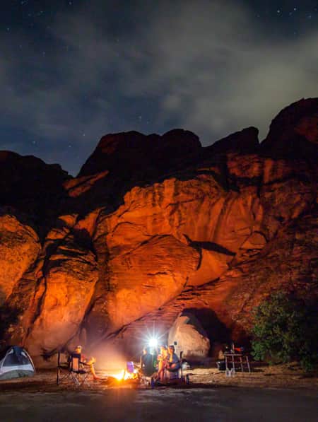 People sitting around a campsite at night, surrounded by red rock formations, with the area illuminated by the light of a campfire.