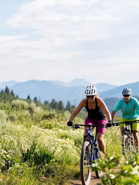Four people cycling on a narrow dirt trail, with green grass and wildflowers on each side.