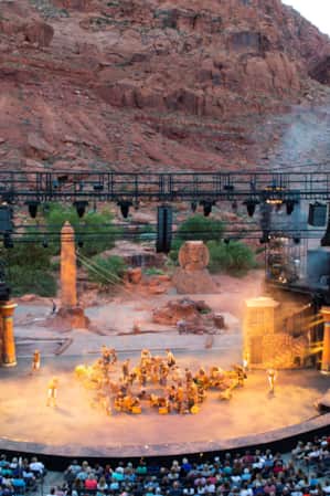 An outdoor amphitheater set against a large red rock, with a crowd of people sitting down and watching a performance.
