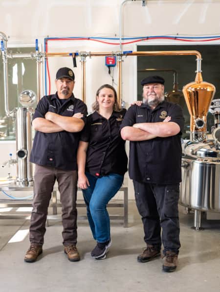 A woman standing between two men, all smiling at the camera with their arms crossed, inside a distillery.