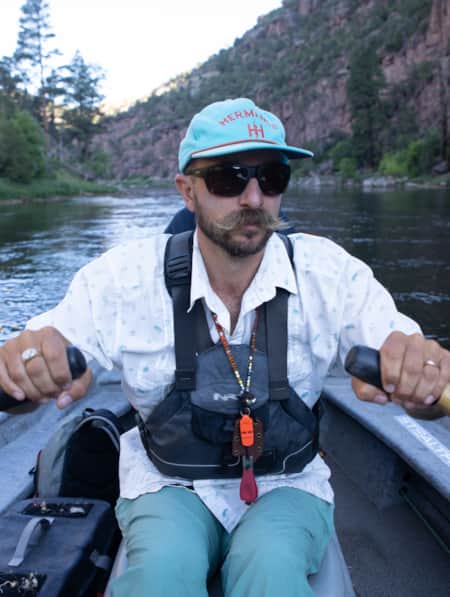 A man rowing a canoe in a river, with fly fishing gear visible in the canoe.