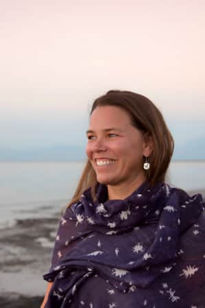 A woman smiling and looking into the distance while wearing a shawl, with a lake in the background.