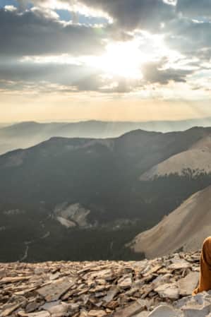 A woman sitting at the summit of a mountain, enjoying the sunrise while drinking a warm beverage and overlooking a view of surrounding mountains.