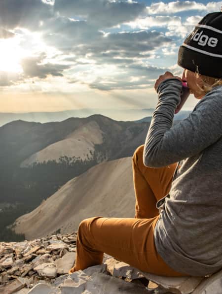 A woman sitting at the summit of a mountain, enjoying the sunrise while drinking a warm beverage and overlooking a view of surrounding mountains.