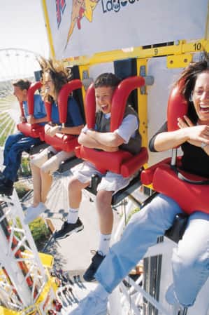 Four people seated at the top of a roller coaster, smiling and yelling as they hold onto the shoulder restraints.