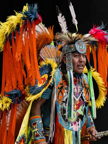 A man dressed in traditional Navajo Hoop dancing attire, featuring orange and yellow feather wings, a feathered headpiece, and a turquoise chest covering with traditional patterns.