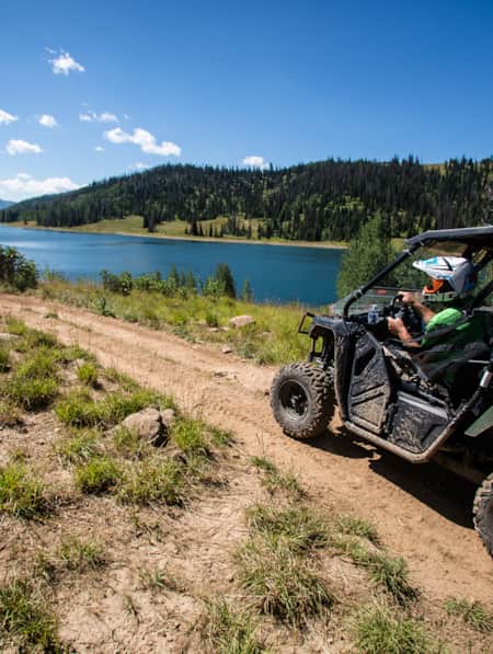 Two ATVs drive along a dirt path beside a lake.